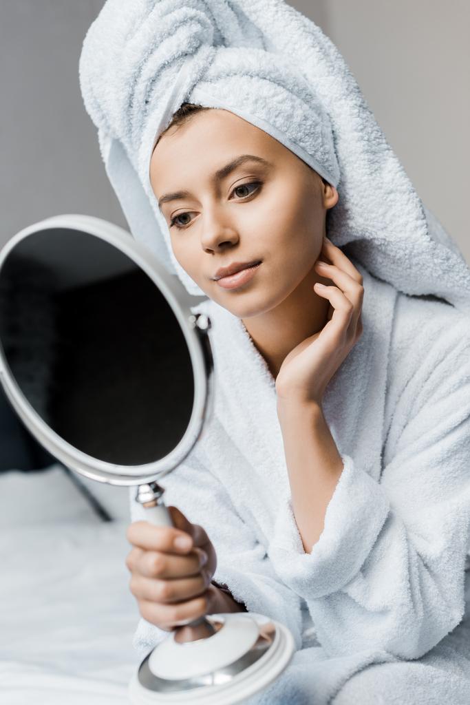 schöne Frau in weißem Bademantel und Handtuch, die ihre perfekte Haut im Spiegel betrachtet   - Foto, Bild