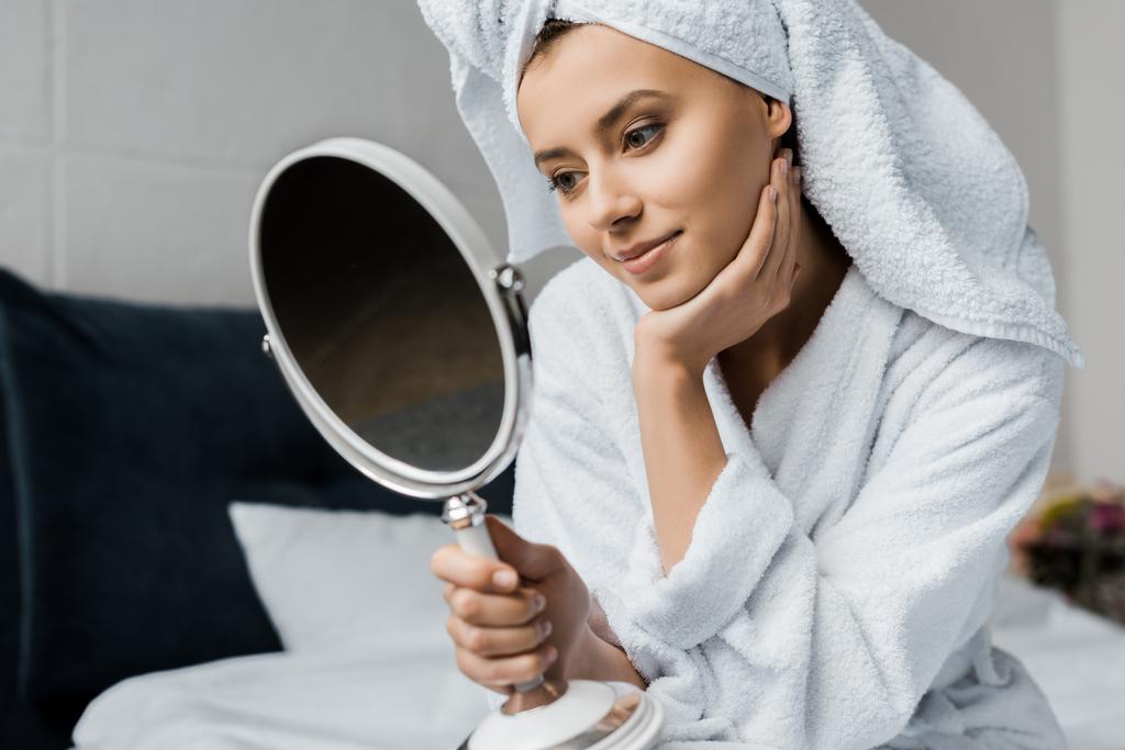 красивая улыбающаяся девушка в белом полотенце смотрит на свое лицо в зеркале
    - Фото, изображение