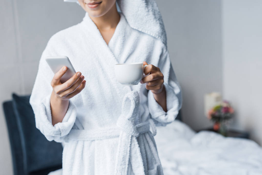 abgeschnittene Ansicht einer Frau im Bademantel, die morgens eine Tasse Kaffee hält und ihr Smartphone im Schlafzimmer benutzt - Foto, Bild