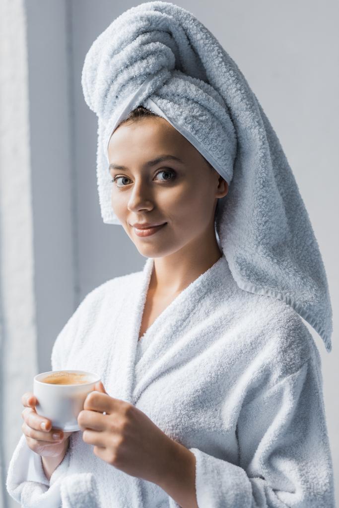 Belle fille en peignoir et serviette blanche sur la tête tenant tasse de café et regardant la caméra
 - Photo, image