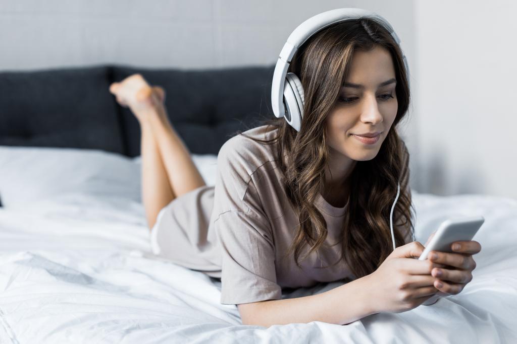 όμορφη μελαχρινή κοπέλα ακούτε μουσική με ακουστικά και smartphone ενώ ξαπλωμένος στο κρεβάτι - Φωτογραφία, εικόνα