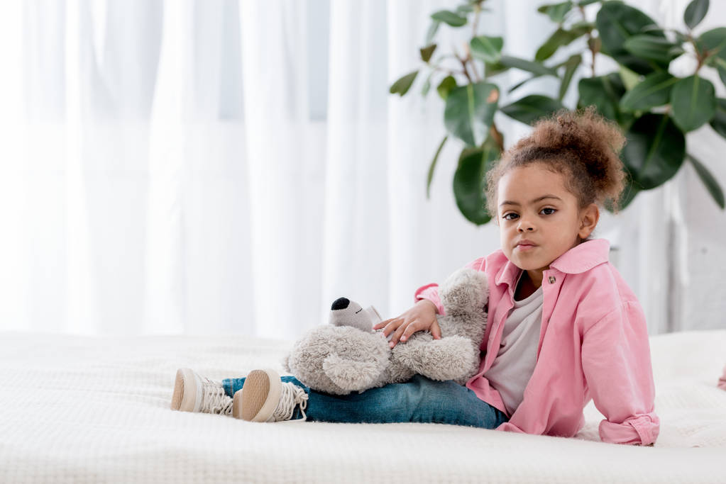 aufgebrachte afrikanisch-amerikanische Kind sitzt auf dem Bett mit Teddybär auf den Knien - Foto, Bild
