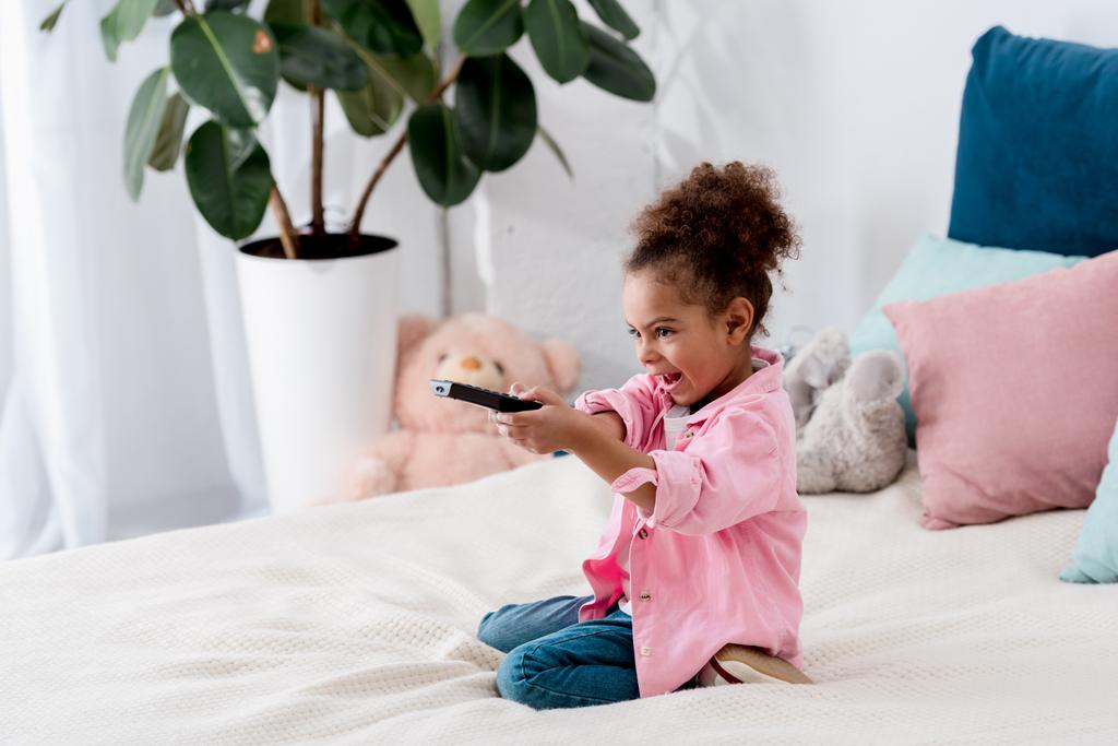 Emocional niño afroamericano sentado en la cama con control remoto de televisión y cambiar los canales
 - Foto, imagen