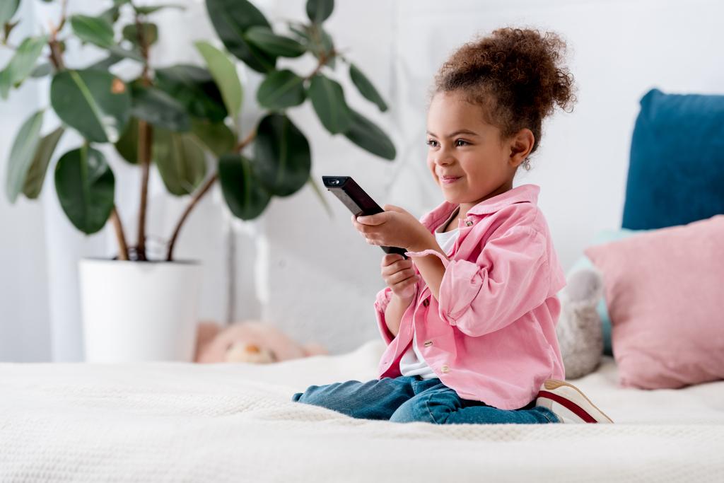 Enfant afro-américain excité assis sur le lit avec télécommande tv
 - Photo, image