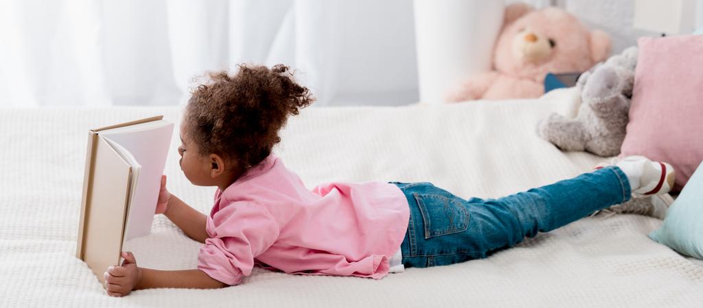 Adorable enfant afro-américain couché sur le lit et lisant un livre
  - Photo, image