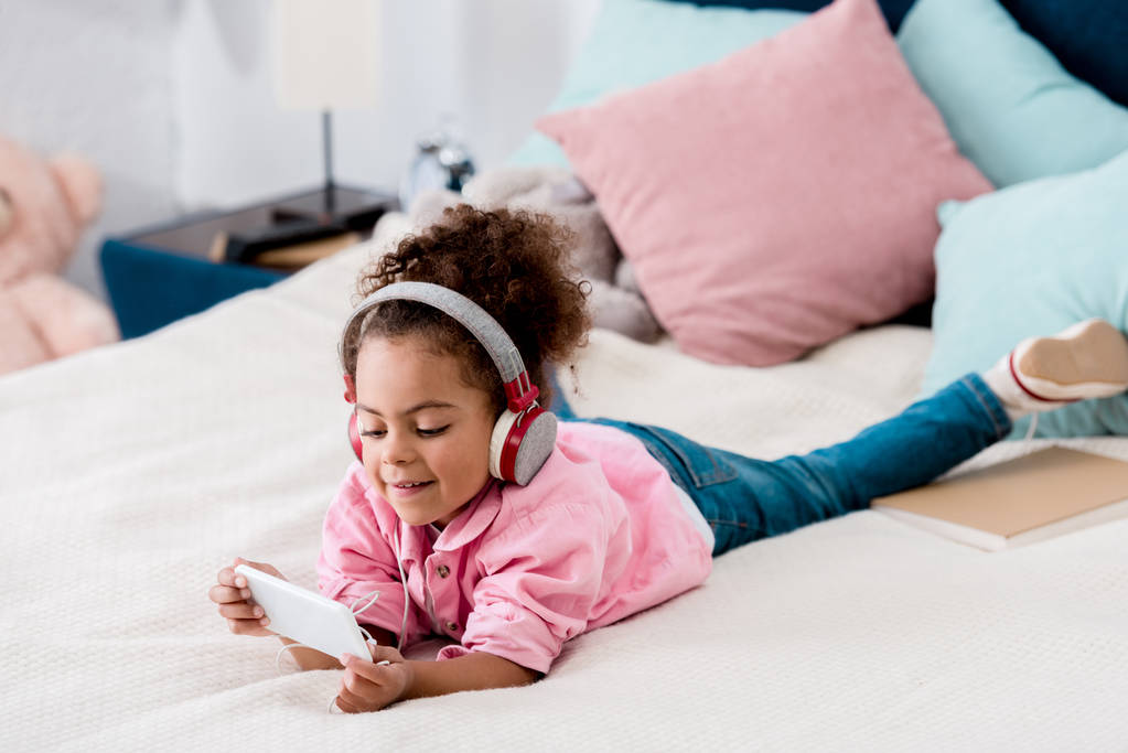 かわいいアフリカ系アメリカ人の子、ベッドの上に横たわるとヘッドホンをスマート フォンで音楽を聴く - 写真・画像