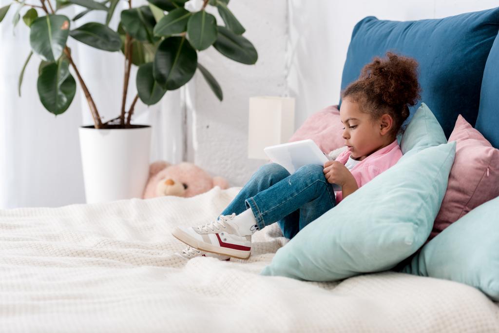 niño afroamericano rizado sentado en la cama con tableta digital
 - Foto, Imagen