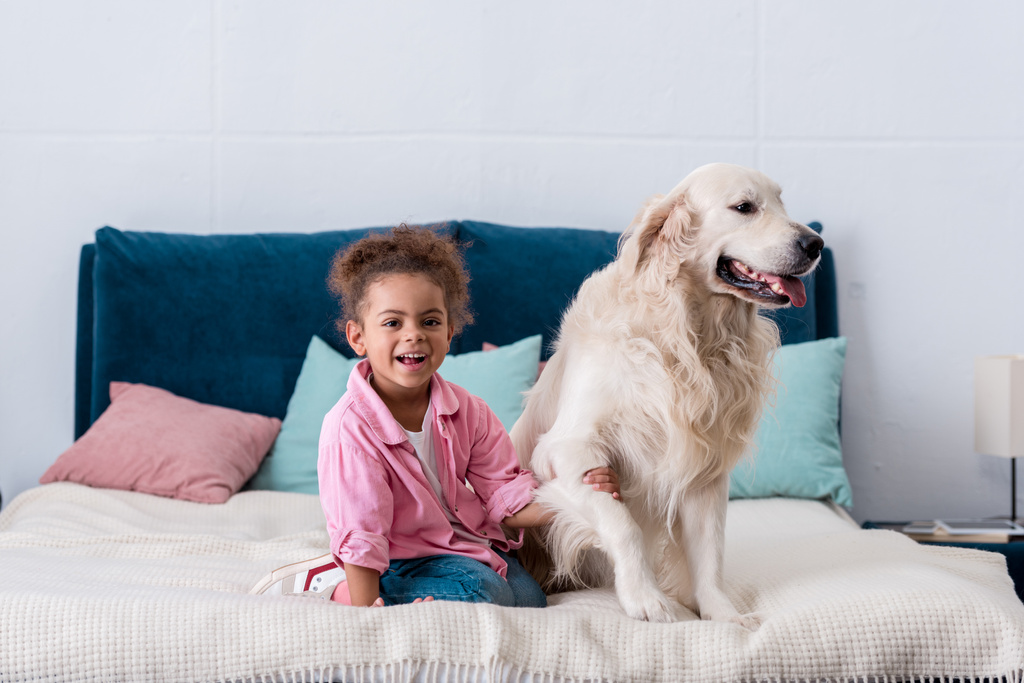 Lachende Afro-Amerikaanse kind zittend op het bed en het bedrijf hond poot - Foto, afbeelding