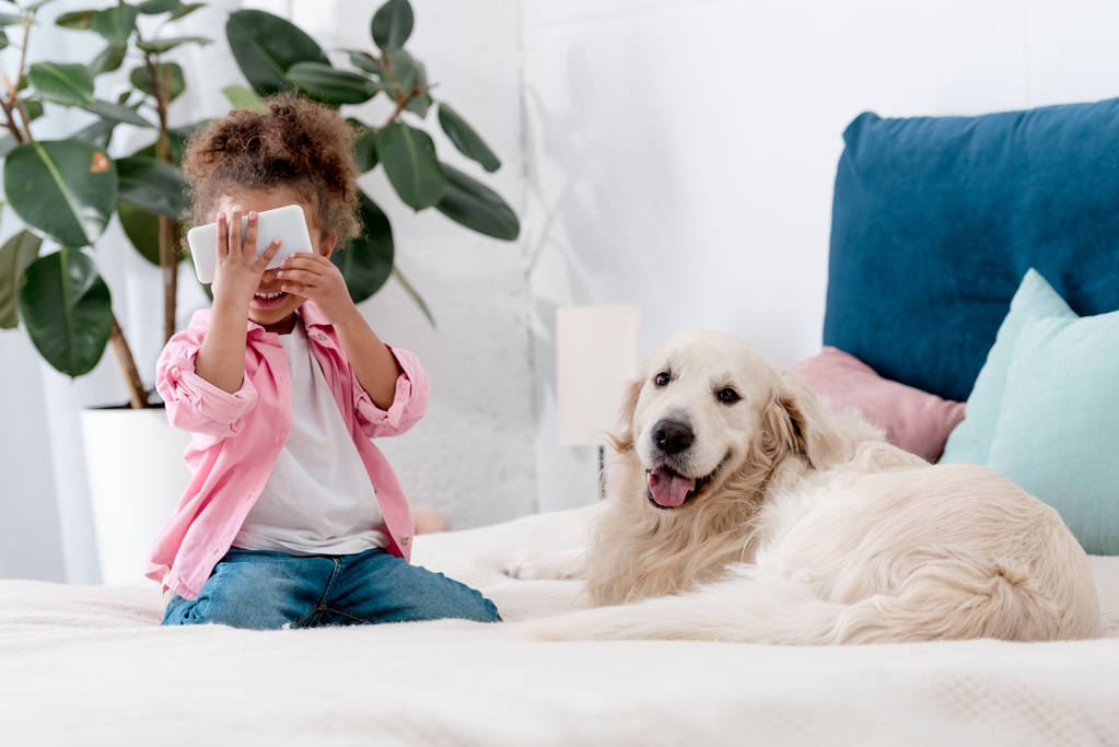 cute african american child sitting on the bed with retriever and holding smartphone near face - Photo, Image