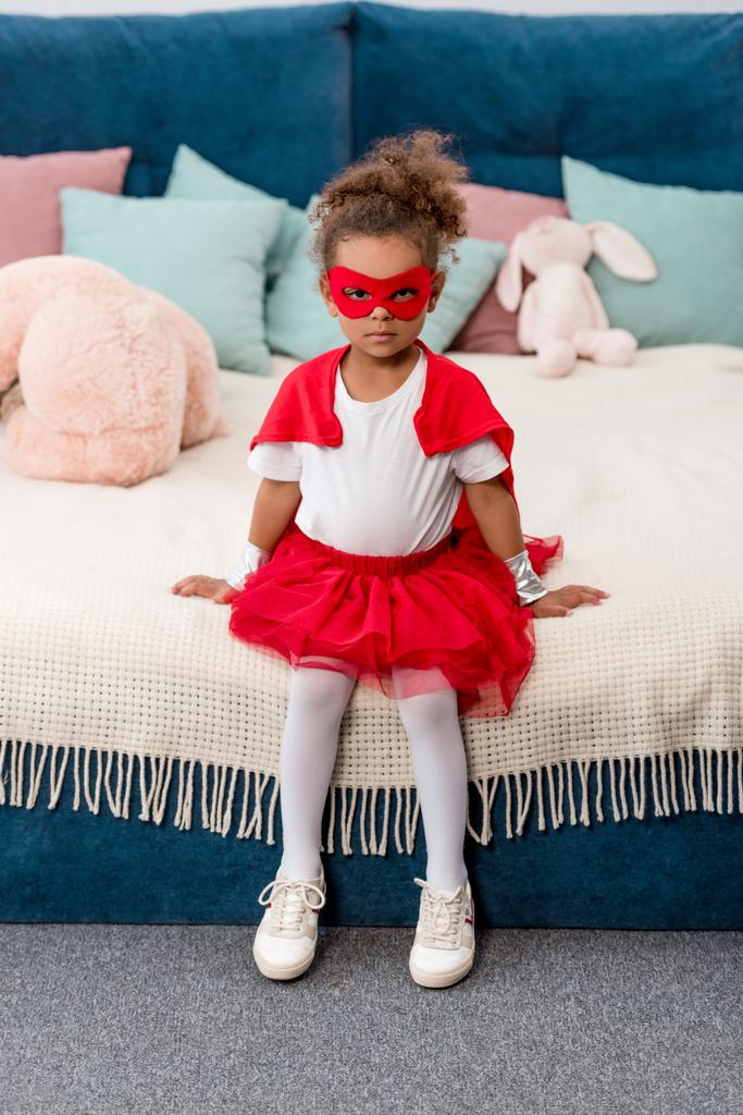 Sevimli küçük Afro-Amerikan çocuk kırmızı süper kahraman kostümü ve yatağa oturmuş maskesi - Fotoğraf, Görsel