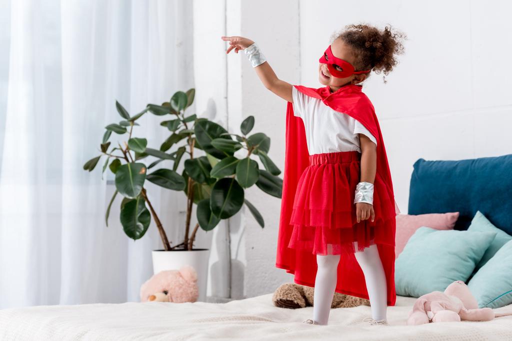 Adorable little african american child in red superhero costume gesturing while standing on bed - Photo, Image