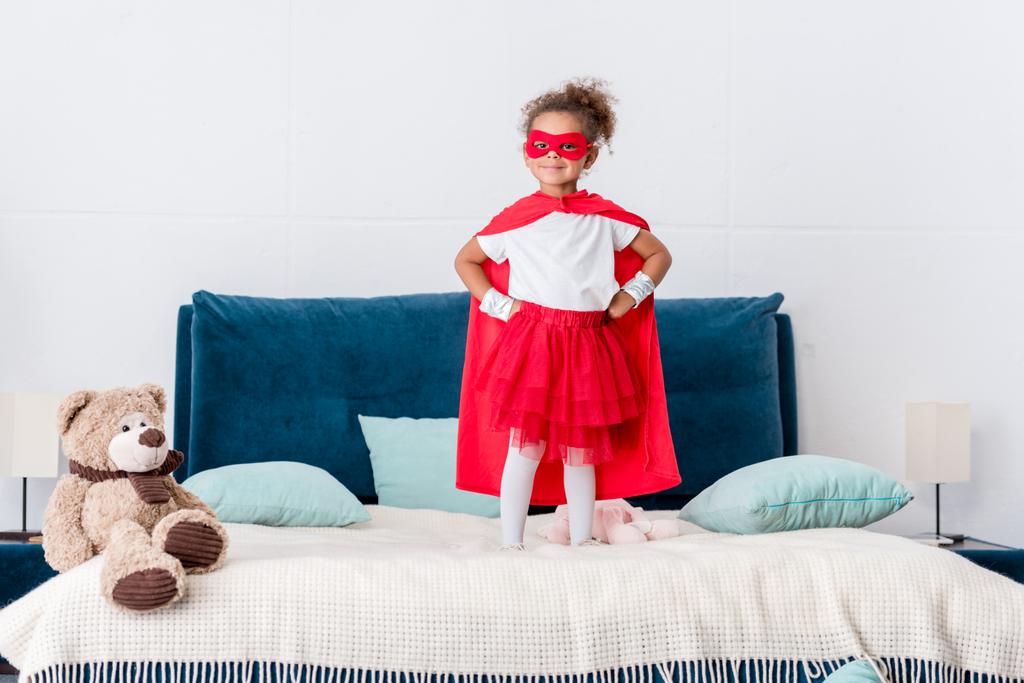 маленька афроамериканська дитина в червоному костюмі супергероя і маска з руками на стегнах, стоячи на ліжку
  - Фото, зображення