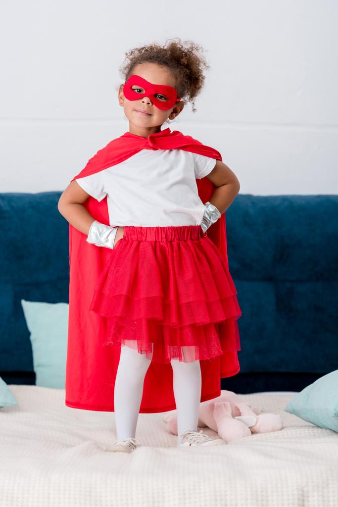 Şirin Afro-Amerikan çocuk kırmızı süper kahraman kostümü ile kalçalarına yatakta ayakta eller  - Fotoğraf, Görsel