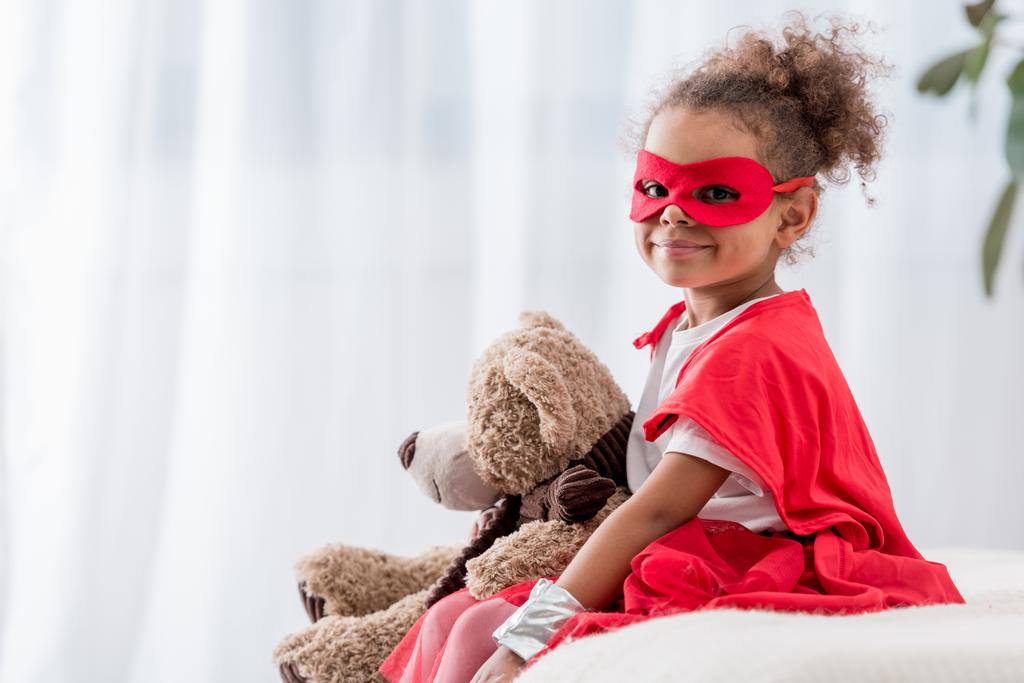 Adorável pequena criança afro-americana em traje de super-herói e máscara com ursinho sorrindo para a câmera
 - Foto, Imagem
