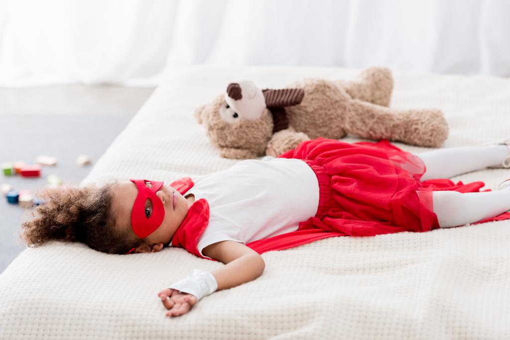 Cute little african american child in superhero costume and mask lying on bed with teddy bear - Photo, Image