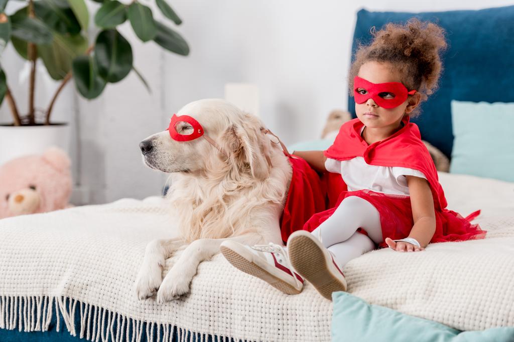 Adorable niño afroamericano con perro en trajes de superhéroe sentado en la cama
 - Foto, Imagen