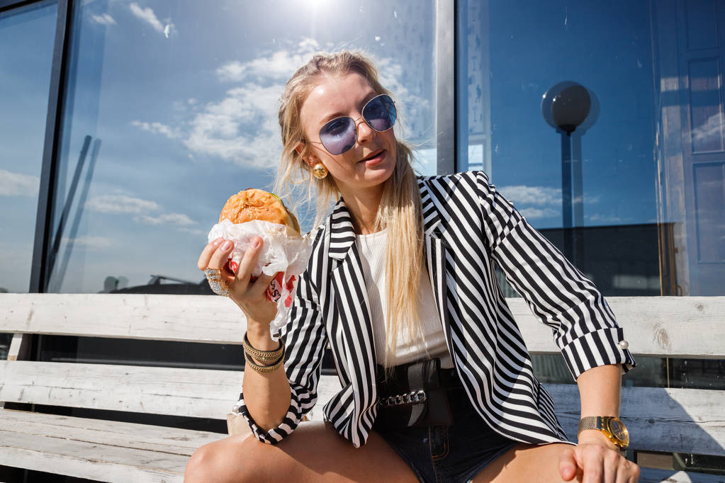 Portrét krásnou blondýnku, nosí brýle, Pruhované sako a bílé tričko jíst sendvič venku - Fotografie, Obrázek