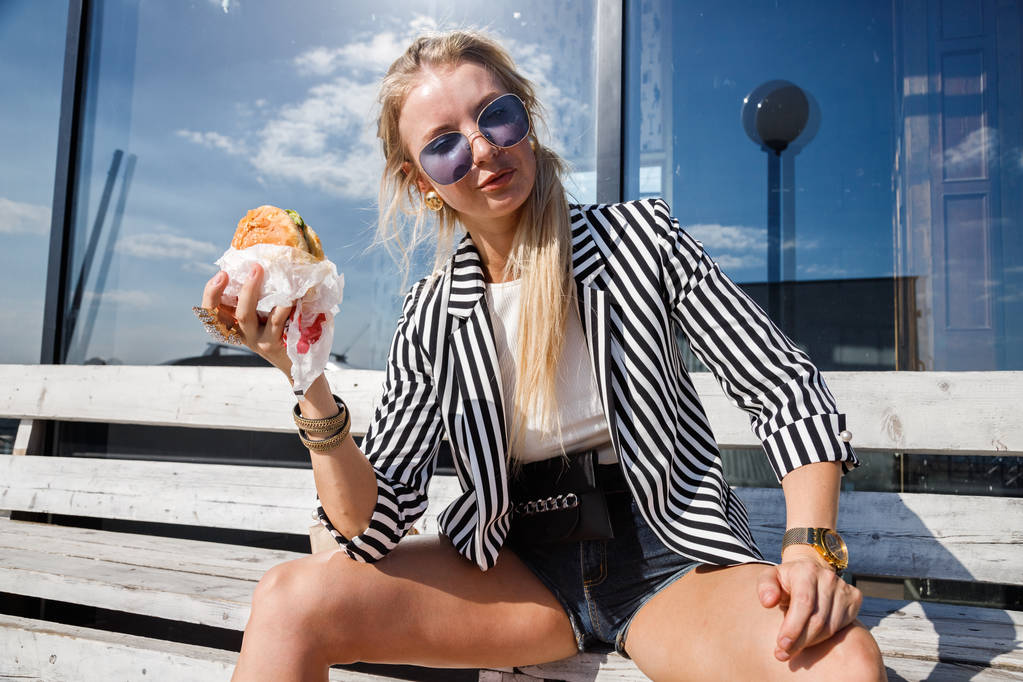 Portret van mooie blonde meisje dragen zonnebril, gestreepte jas en witte t-shirt eten sandwich buitenshuis - Foto, afbeelding