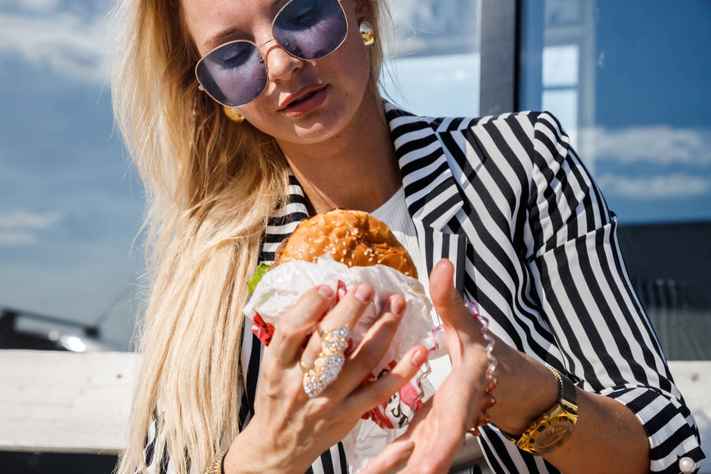 Portrait of lovely blonde girl wearing sunglasses, striped jacket and white t-shirt eating sandwich outdoors - Photo, Image