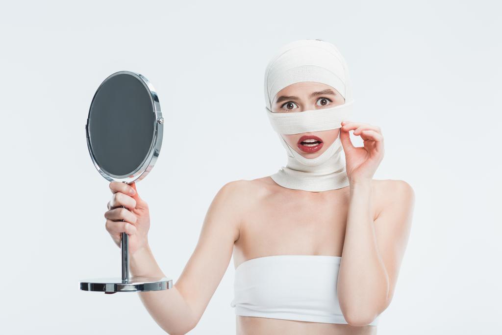 испуганная женщина с бинтами над головой, держащая зеркало, изолированное на белом
 - Фото, изображение