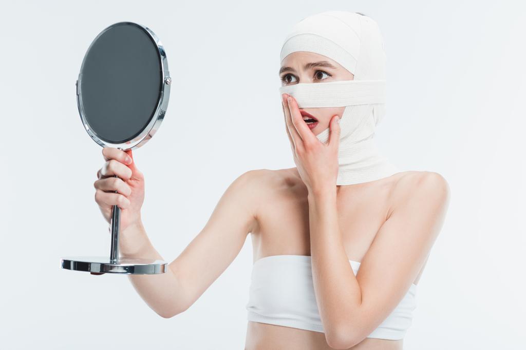 mujer conmocionada con vendajes sobre la cabeza mirando el espejo aislado en blanco
 - Foto, imagen