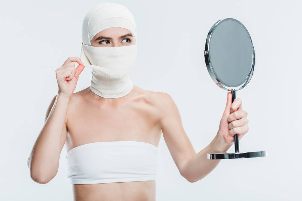 woman with bandages after plastic surgery on face looking at mirror isolated on white - Photo, Image