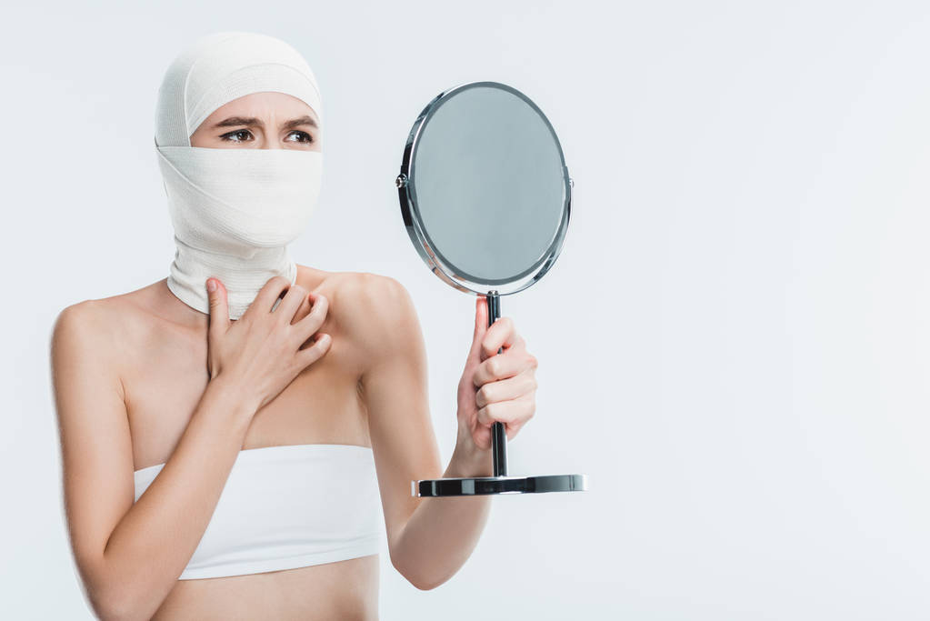 verängstigte Frau nach Schönheitsoperation im Gesicht mit Blick auf Spiegel isoliert auf Weiß - Foto, Bild