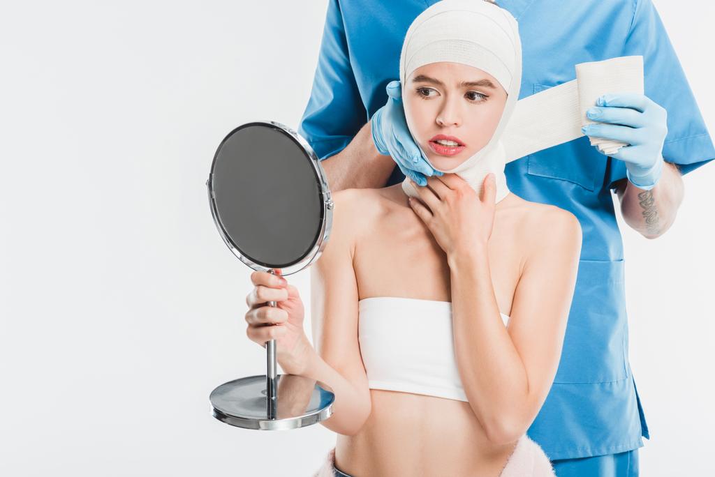 Χειρουργός στα γάντια δέσει επάνω πρόσωπο με επίδεσμο μετά από πλαστικές επεμβάσεις ενώ γυναίκα κοιτάζοντας καθρέφτη που απομονώνονται σε λευκό - Φωτογραφία, εικόνα