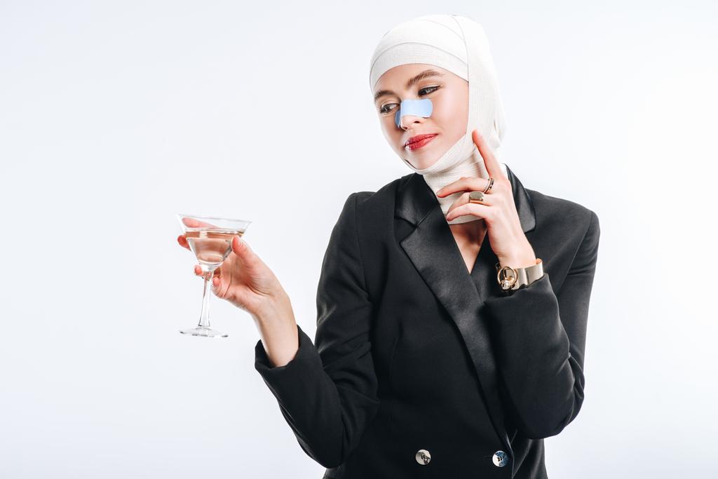 стильна усміхнена жінка з бинтами над головою після пластичної хірургії тримає скло з коктейлем ізольовано на білому
 - Фото, зображення