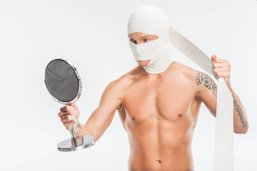 naked man taking off bandages from head after plastic surgery and looking at mirror isolated on white - Photo, Image