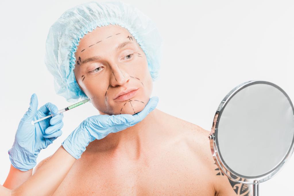 Руки хирурга держат шприц, в то время как человек смотрит на зеркало, изолированное на белом
 - Фото, изображение