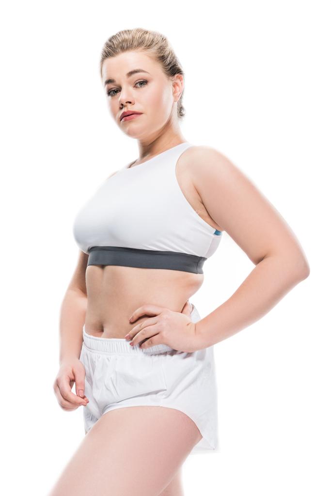 atractiva chica con sobrepeso en ropa deportiva de pie con la mano en la cintura y mirando a la cámara aislada en blanco
 - Foto, Imagen