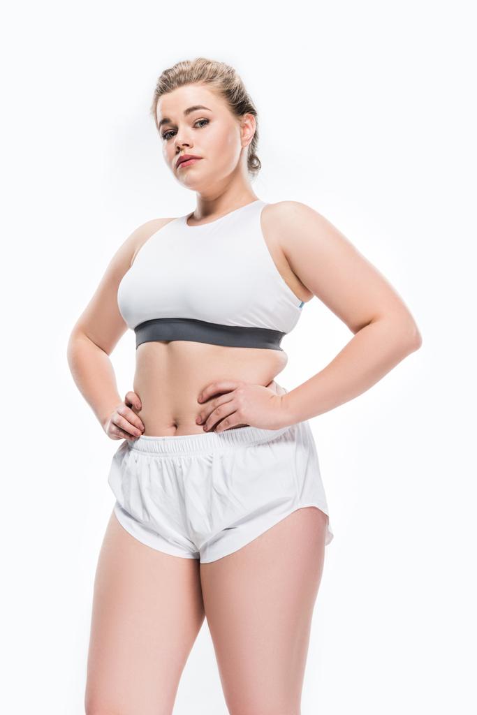 Niedrigwinkelaufnahme einer jungen übergewichtigen Frau in Sportbekleidung, die mit den Händen auf der Taille steht und isoliert auf weiß in die Kamera blickt  - Foto, Bild