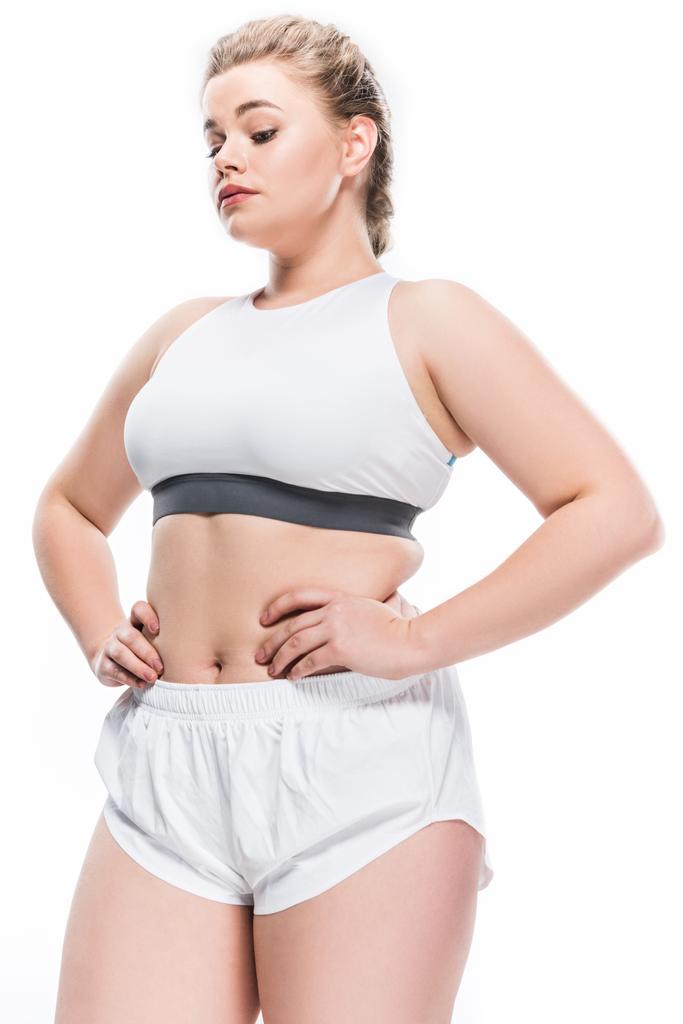 Niedrigwinkel-Ansicht einer jungen übergewichtigen Frau in Sportkleidung, die mit den Händen auf der Taille steht, isoliert auf weiß  - Foto, Bild