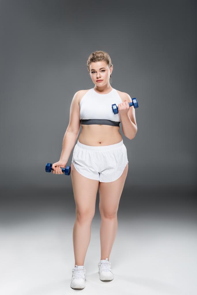 junge übergroße Frau in Sportbekleidung, die mit Kurzhanteln trainiert und in die Kamera auf grau blickt  - Foto, Bild