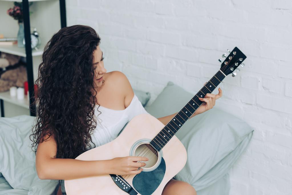 усміхнена кучерява дівчина грає на акустичній гітарі в спальні вдома
 - Фото, зображення