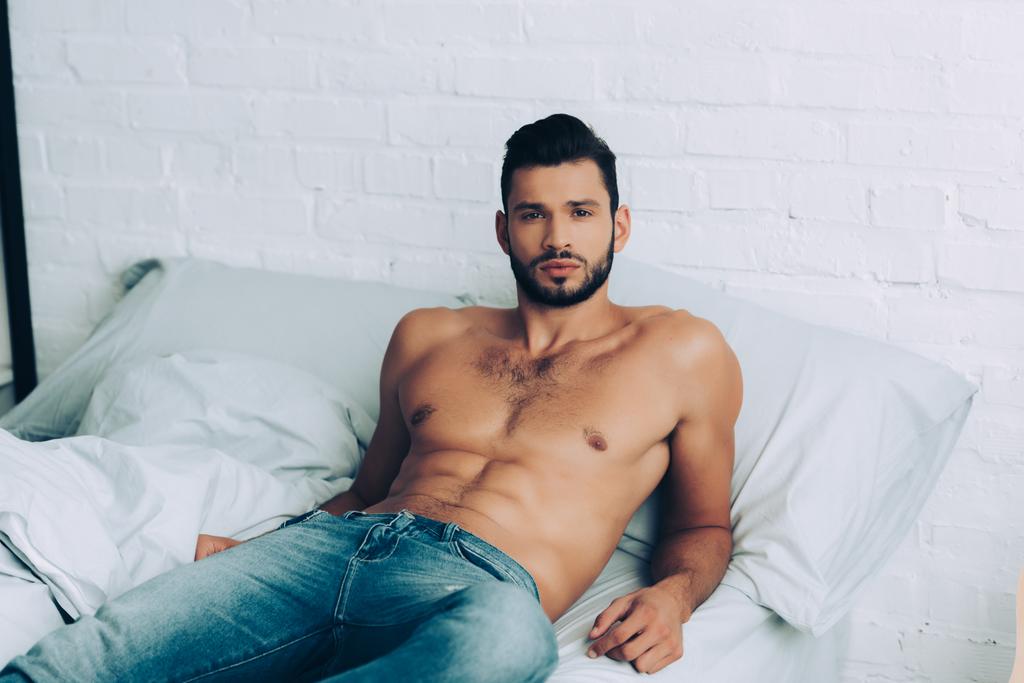 красивый мускулистый мужчина без рубашки в джинсах позирует на кровати дома
 - Фото, изображение