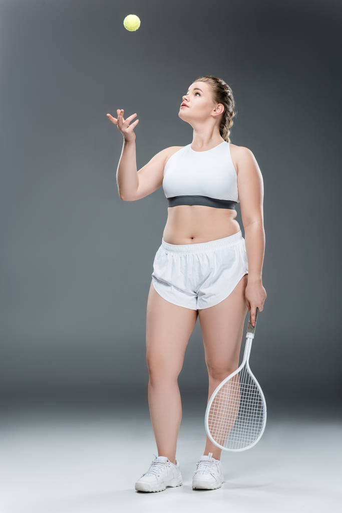Młoda kobieta z nadwagą przytrzymanie rakieta tenisowa i rzucając piłkę na szary - Zdjęcie, obraz