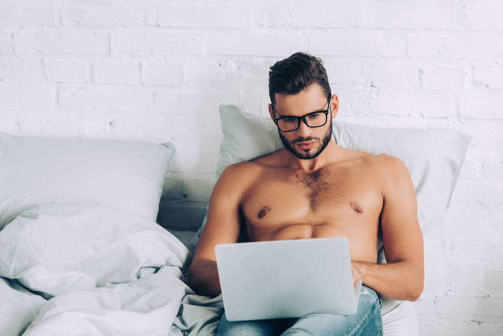 красивый мужчина без рубашки фрилансер с мускулистым туловищем работает на ноутбуке в постели дома
 - Фото, изображение