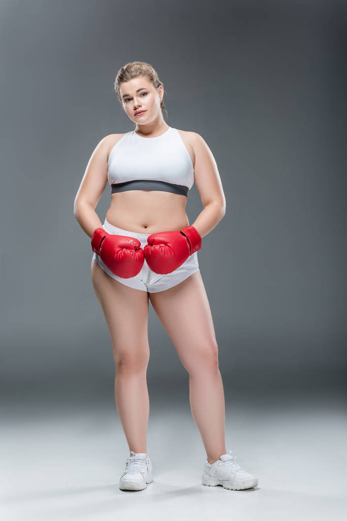 full length view of young overweight woman in sportswear and boxing gloves looking at camera on grey - Photo, Image