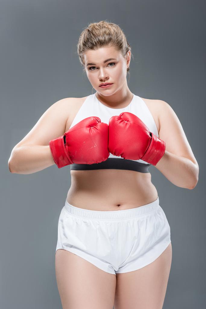 kaunis nuori ylipainoinen nainen urheiluvaatteissa ja nyrkkeilyhanskat katsoen kameran eristetty harmaa
 - Valokuva, kuva
