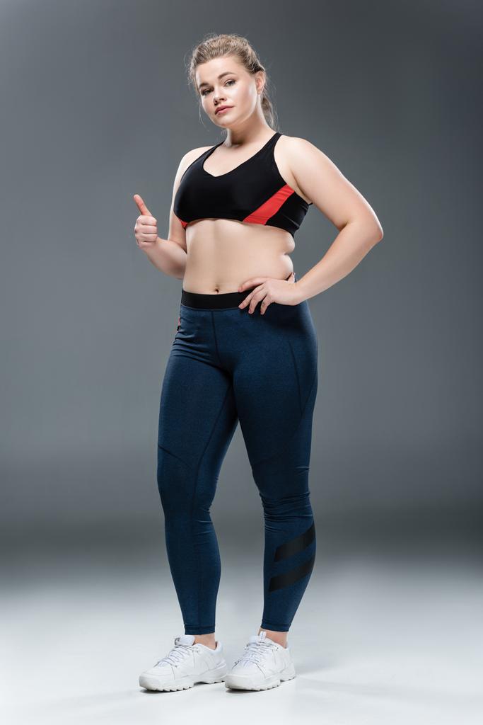 beautiful overweight girl in sportswear showing thumb up and looking at camera on grey - Photo, Image