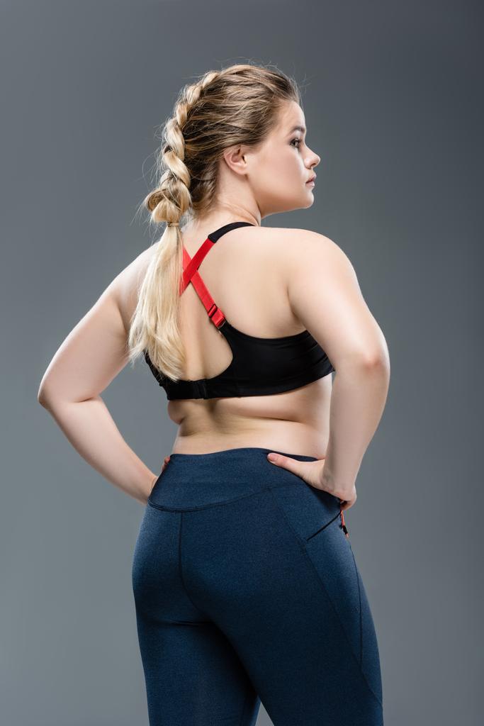 вид ззаду молодої надмірної ваги жінки в спортивному одязі, що стоїть з руками на талії ізольовано на сірому
 - Фото, зображення