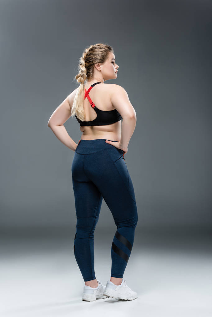 visão traseira da jovem mulher com sobrepeso em sportswear de pé com as mãos na cintura em cinza
 - Foto, Imagem
