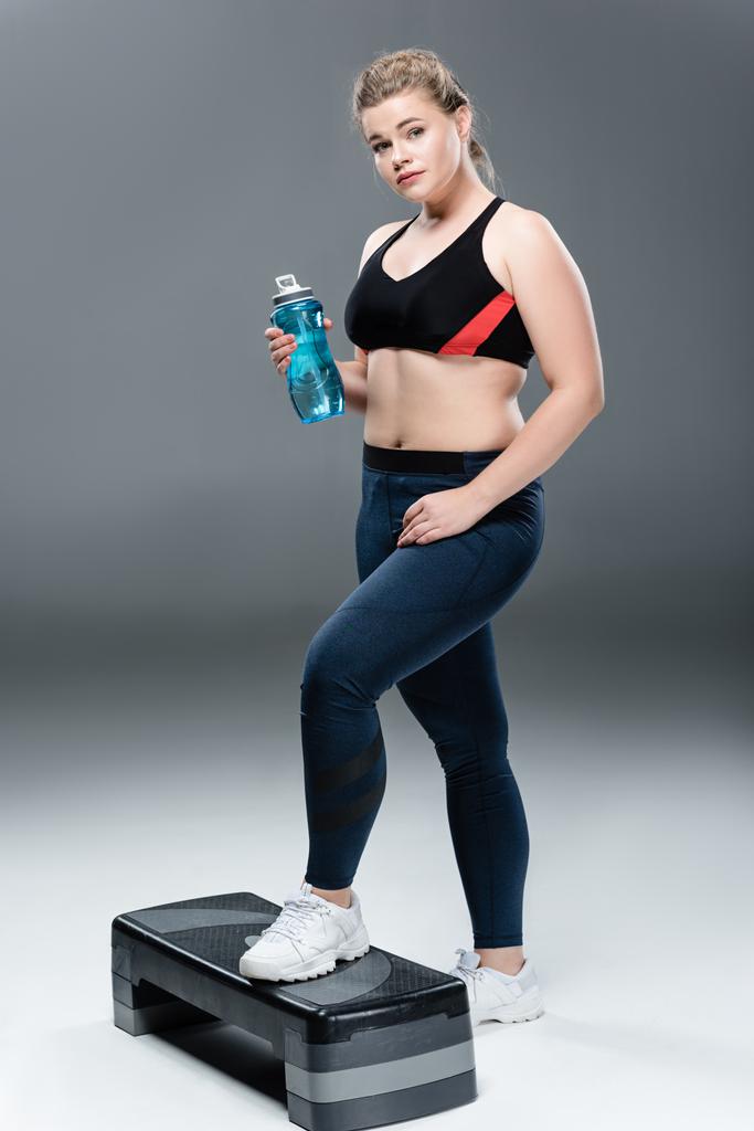 giovane donna oversize in abbigliamento sportivo in possesso di bottiglia d'acqua e in piedi su piattaforma gradino su grigio
  - Foto, immagini