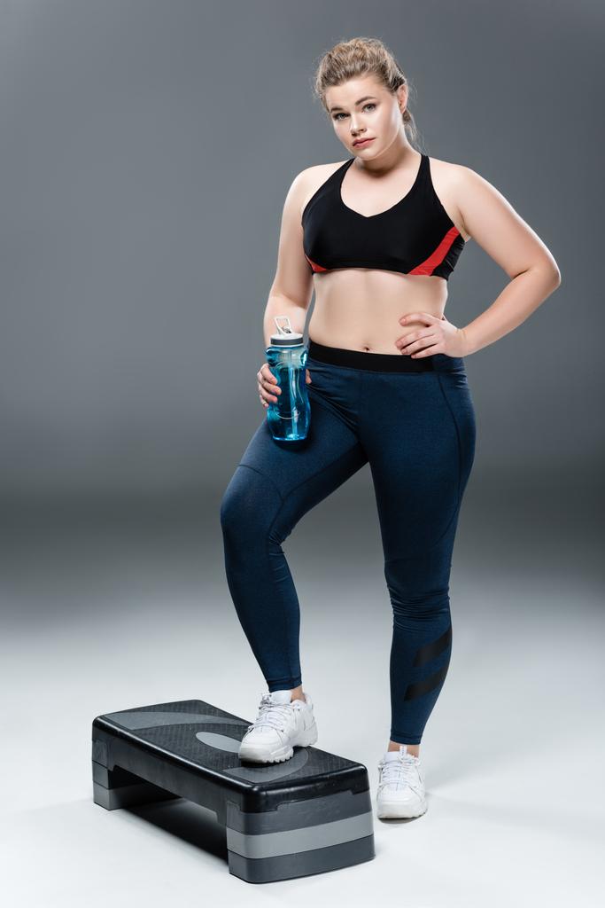 νεαρή γυναίκα υπέρβαροι σε αθλητικά ρούχα κρατώντας το μπουκάλι νερό και να στέκεται στην πλατφόρμα βήμα σε γκρι  - Φωτογραφία, εικόνα