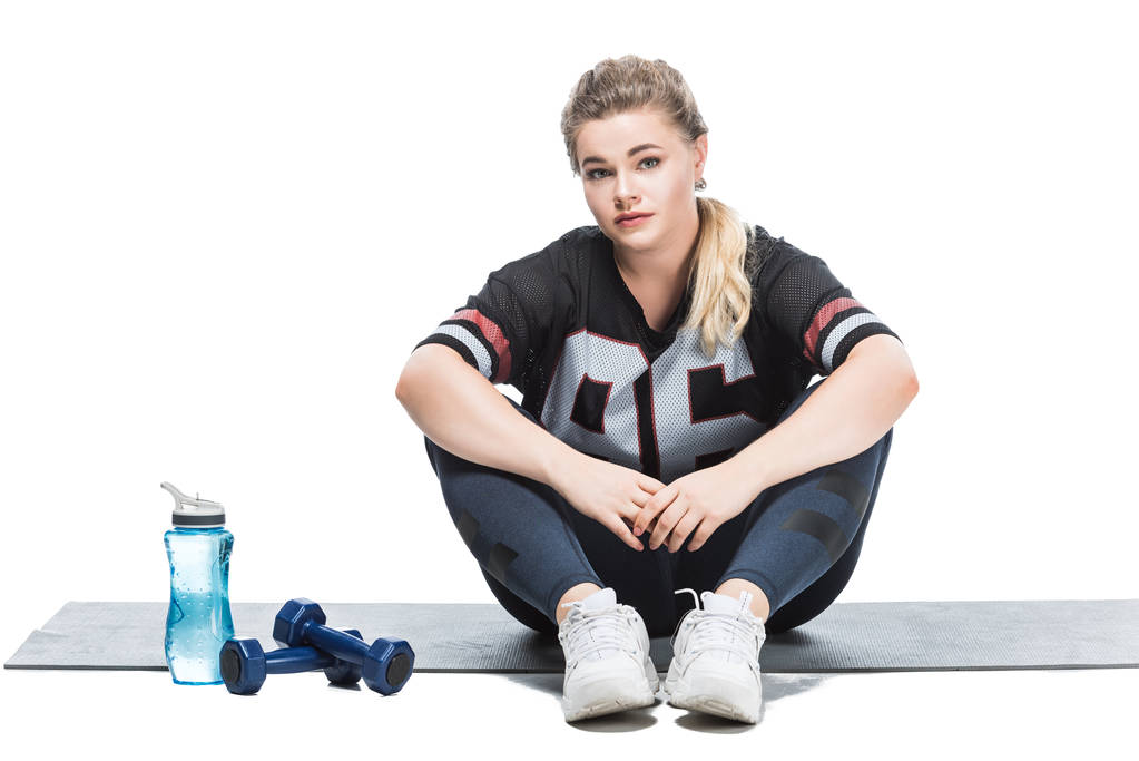 tamanho jovem mais mulher em sportswear sentado no tapete de ioga e olhando para a câmera isolada no branco
   - Foto, Imagem
