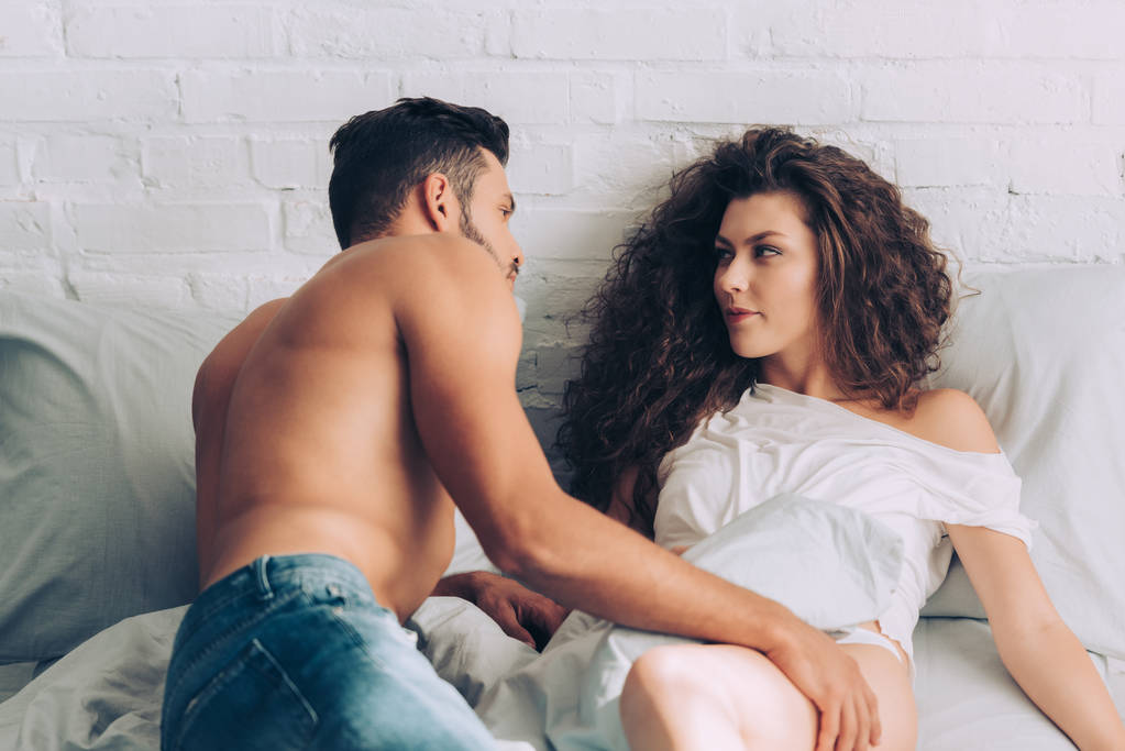 handsome muscular man in jeans and his curly attractive girlfriend on bed at home - Photo, Image