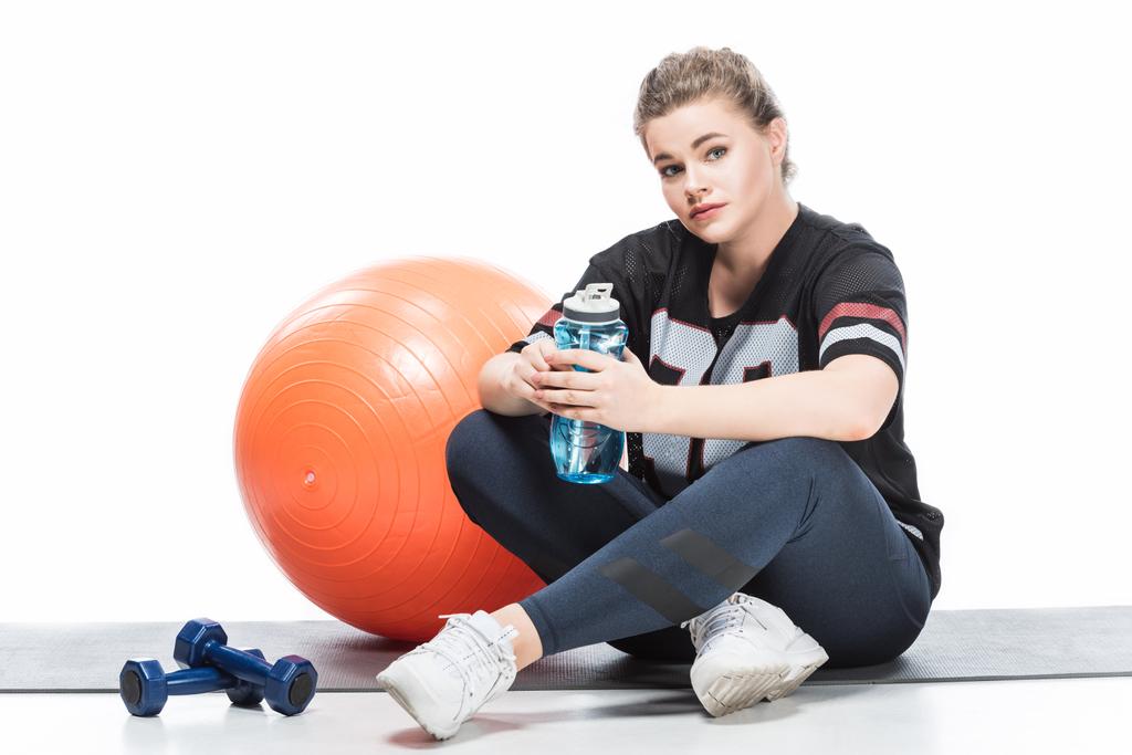 young overweight woman in sportswear holding bottle of water and looking at camera while sitting on yoga mat isolated on white - Photo, Image