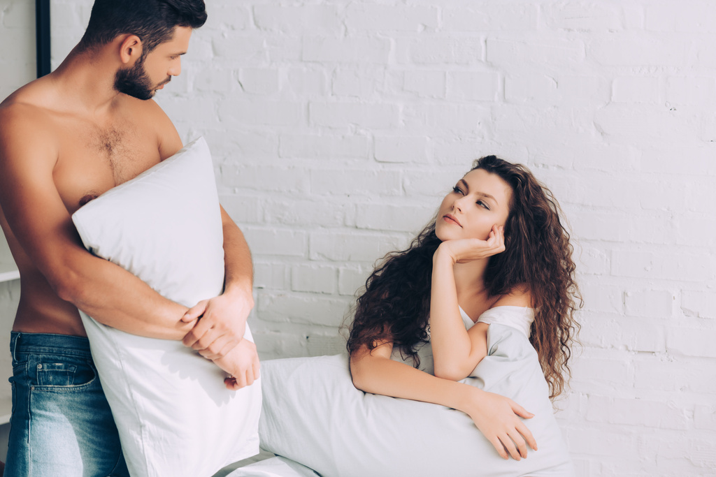 красивый мускулистый мужчина в джинсах держит подушку и разговаривает с девушкой в спальне дома
 - Фото, изображение